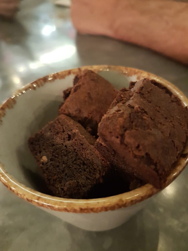 A small ceramic bowl filled with several thick, square pieces of rich, dark chocolate brownie. The brownies appear dense and fudgy, with slightly crumbly edges. The background is softly blurred, highlighting the brownies.