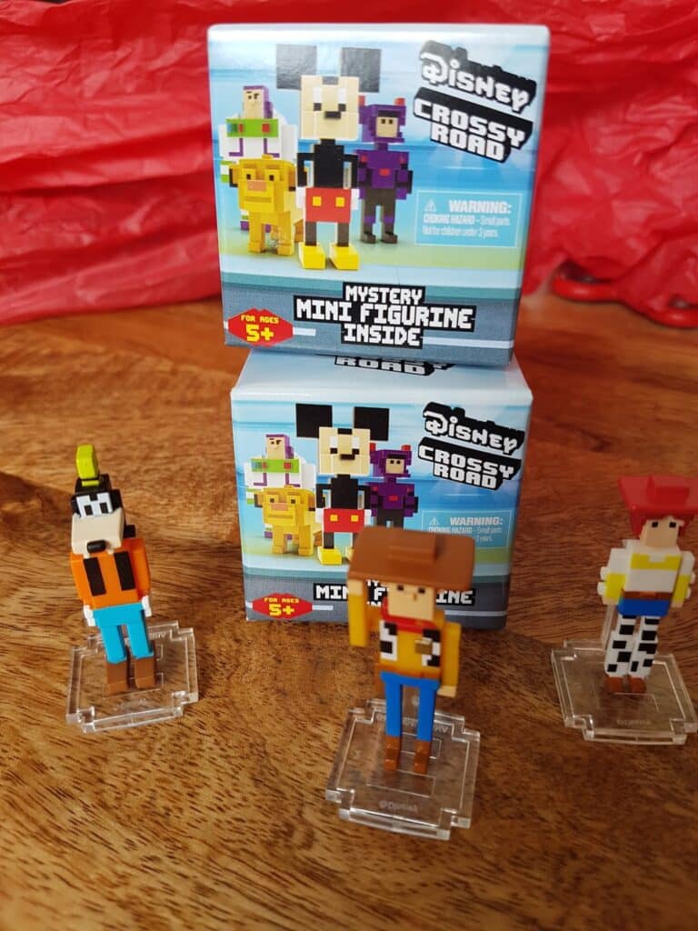 Three pixelated Disney character figurines—Goofy, Woody, and Jessie—stand on clear bases in front of two Disney Crossy Road “Mystery Mini Figurine” boxes, set on a wooden surface with red crinkled paper in the background.