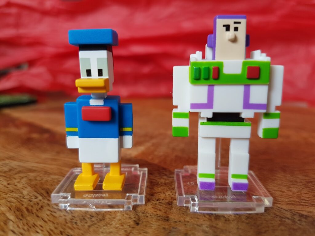Two blocky, pixelated toy figures stand on clear bases. The left figure is Donald Duck, wearing a blue sailor suit and hat. The right figure is Buzz Lightyear, in a white spacesuit with green and purple accents. Both are on a wooden surface with a red background.