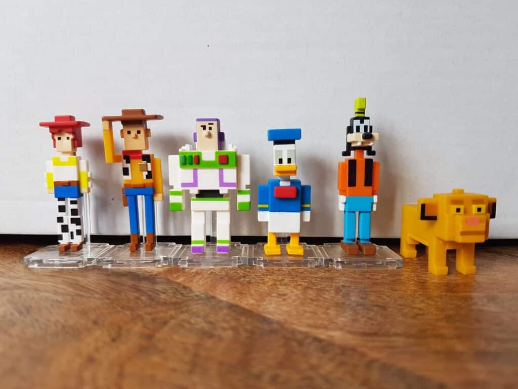 Six pixel-style figurines of Disney characters stand in a row on clear bases: Jessie, Woody, Buzz Lightyear, Donald Duck, Goofy, and Dug the dog, all set against a plain white background and brown wooden surface.