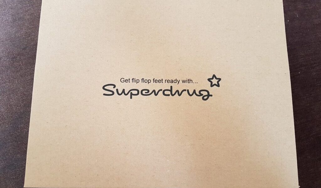 A plain light brown cardboard box sits on a dark surface. Centred on the lid, black text reads: "Get flip flop feet ready with… Superdrug," with a small star symbol next to the brand name.