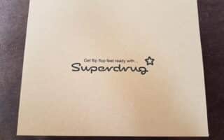 A plain light brown cardboard box sits on a dark surface. Centred on the lid, black text reads: "Get flip flop feet ready with… Superdrug," with a small star symbol next to the brand name.