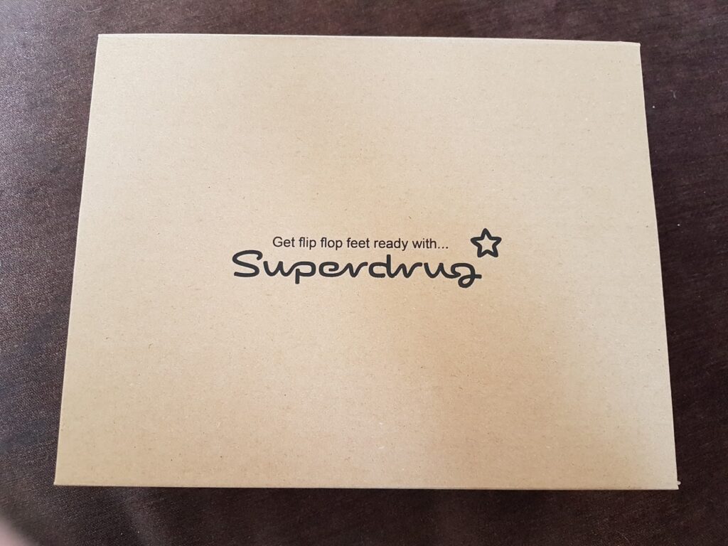 A plain brown cardboard box lies on a dark surface. The top of the box features black text that reads, "Get flip flop feet ready with... Superdrug," with a small star icon near the end of the sentence.