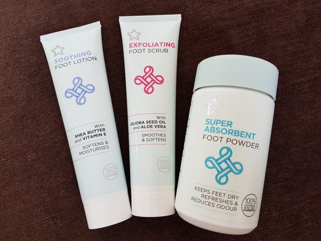 Three foot care products on a brown surface: a tube of soothing foot lotion, a tube of exfoliating foot scrub, and a container of super absorbent foot powder. All packaging is pale with blue text and abstract logo designs.