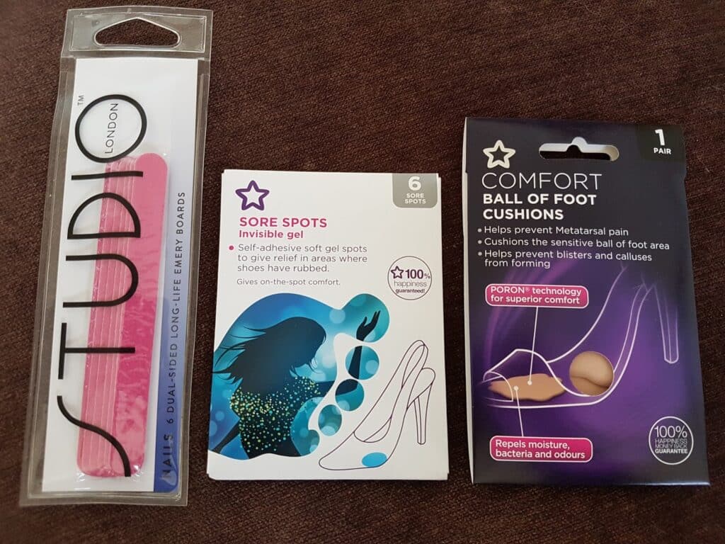 Three products on a brown surface: a pink Studio London nail file in clear packaging, Superdrug Sore Spots Invisible Gel (box with illustration of a woman and high heels), and Comfort Ball of Foot Cushions (box with product image and text).