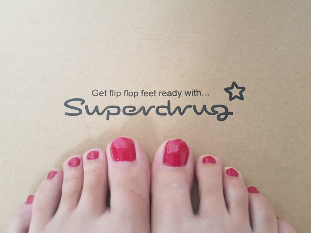 A close-up of toes with bright pink nail varnish on a tan background. Above the toes, text reads: "Get flip flop feet ready with... Superdrug" next to a small star symbol.