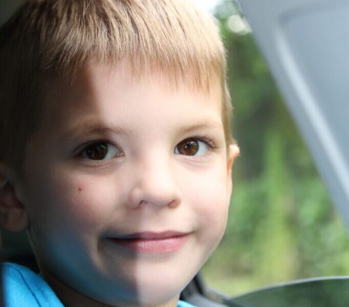 A young boy with short, light brown hair and brown eyes smiles softly whilst sitting by a car window. He wears a blue shirt and holds a green and orange box. Sunlight illuminates his face, and green trees are blurred outside the window.