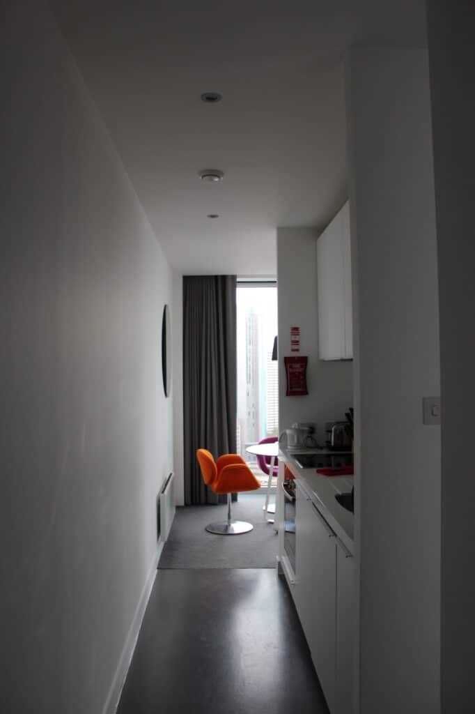 A narrow, modern kitchen with white walls and cupboards leads to a small dining area with a round table and an orange chair by a large window covered by grey curtains. The space is minimally decorated and well-lit by natural light from the window.