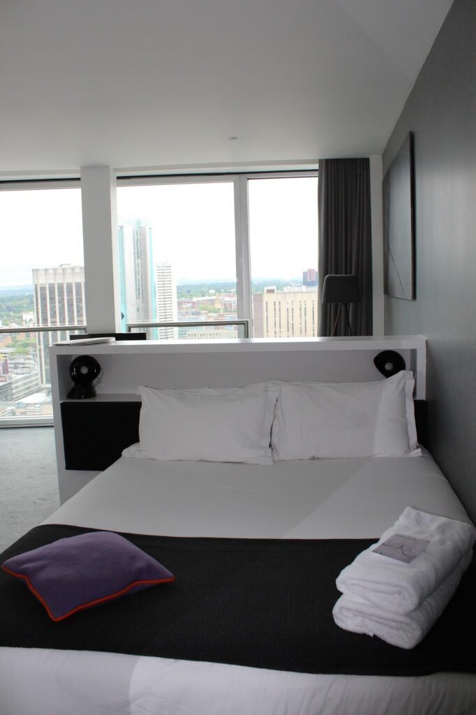 A modern hotel room with a neatly made double bed, white linen, and two pillows. A folded towel and purple-edged blanket are at the foot. Large windows reveal a cityscape with tall buildings. The room has grey walls, minimal décor, and natural light.