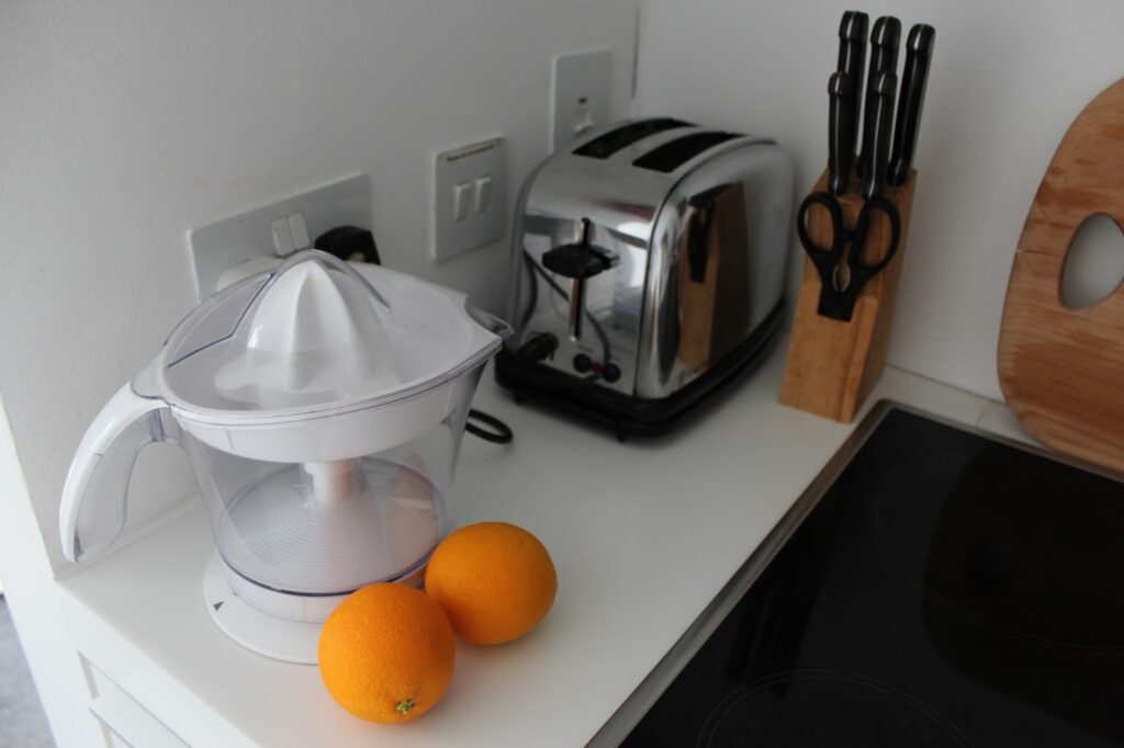 A kitchen worktop with a white citrus juicer, two whole oranges, a shiny silver toaster, a wooden knife block holding knives and scissors, and part of a black hob. The scene is clean and modern with minimal decor.