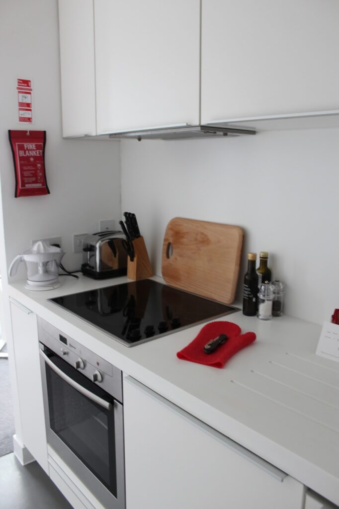 A modern white kitchen with an electric cooker, oven, chopping board, toaster, knife block, citrus juicer, olive oil and vinegar bottles, and a red oven glove on the worktop. A fire blanket is mounted on the wall. Cupboards are above the worktop.