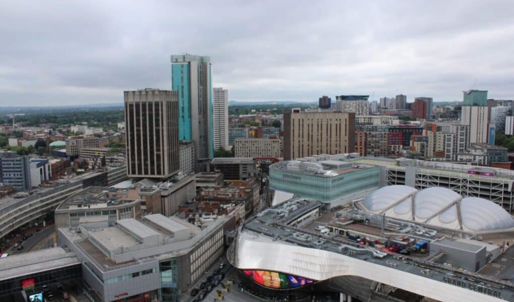 Aerial view of Birmingham city centre featuring modern high-rise buildings, the reflective curved roof of Grand Central shopping centre, and New Street railway station. The sky is overcast, and city streets and varied architecture are visible below.
