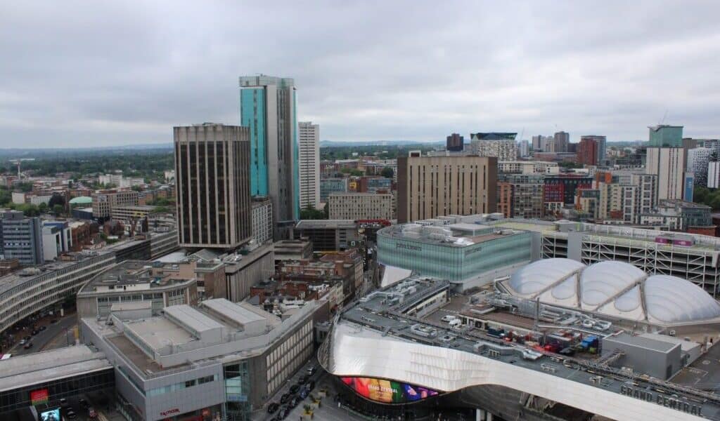 Aerial view of Birmingham city centre featuring modern high-rise buildings, the reflective curved roof of Grand Central shopping centre, and New Street railway station. The sky is overcast, and city streets and varied architecture are visible below.