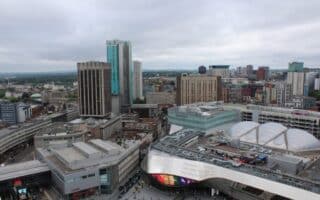Aerial view of Birmingham city centre featuring modern high-rise buildings, the reflective curved roof of Grand Central shopping centre, and New Street railway station. The sky is overcast, and city streets and varied architecture are visible below.