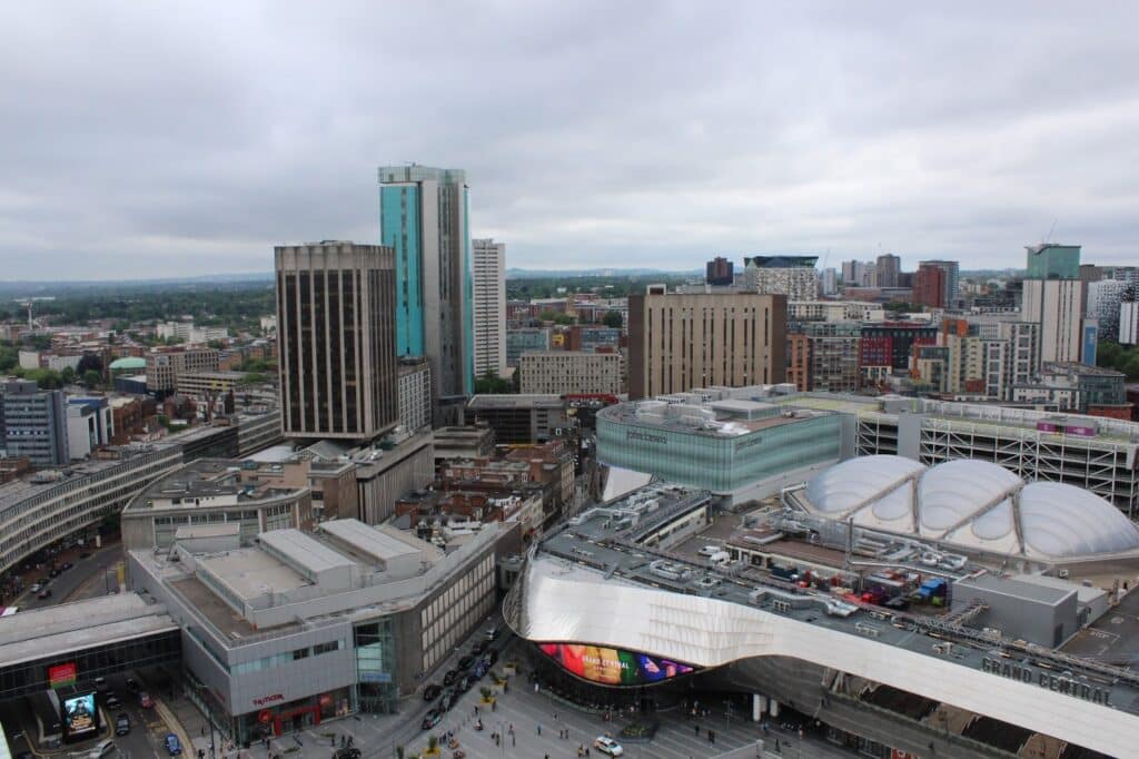 Aerial view of Birmingham city centre, featuring modern tall buildings, the silver curved roof of Grand Central station, glass skyscrapers, and a busy street below with people and vehicles. The sky is overcast with grey clouds.