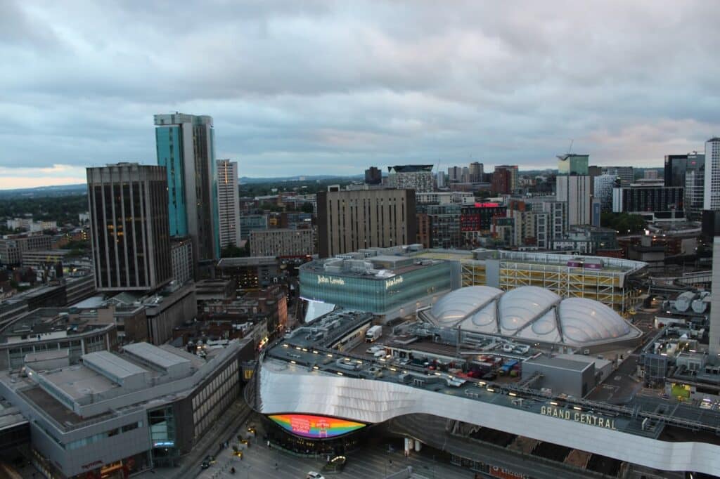 Aerial view of Birmingham city centre showing modern buildings, the Grand Central shopping centre with its curved silver facade, and the adjacent New Street railway station. The sky is cloudy, and city lights are just starting to glow.