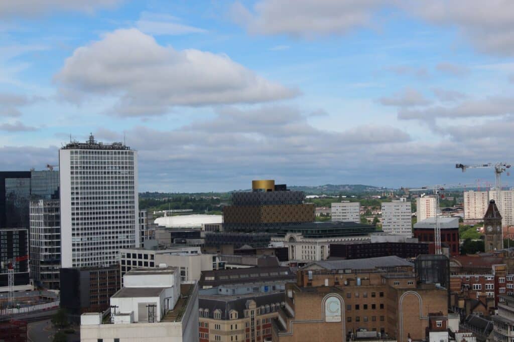A cityscape view of Birmingham, UK, showing modern and historic buildings under a partly cloudy blue sky. The Library of Birmingham, with its distinctive gold and black design, stands out in the centre. Construction cranes and distant green hills are visible in the background.
