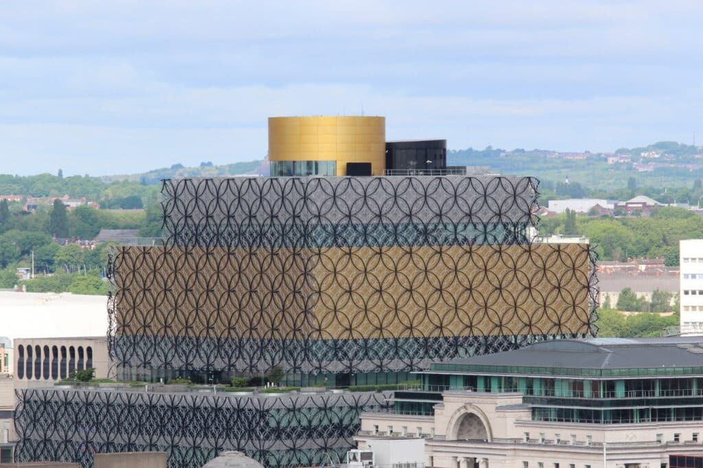 A modern building with three stacked rectangular sections, each with decorative black circular latticework. The middle section is gold, while the others are grey. A round gold structure sits on top. The building is surrounded by greenery and other city buildings.