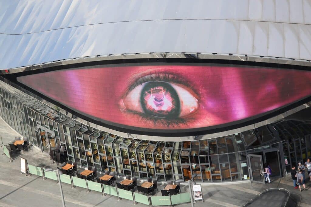 A building with a curved facade features a large digital screen displaying a realistic, detailed human eye. Below are glass windows and outdoor café tables, some with people walking nearby. The building’s silver roof arches above the scene.