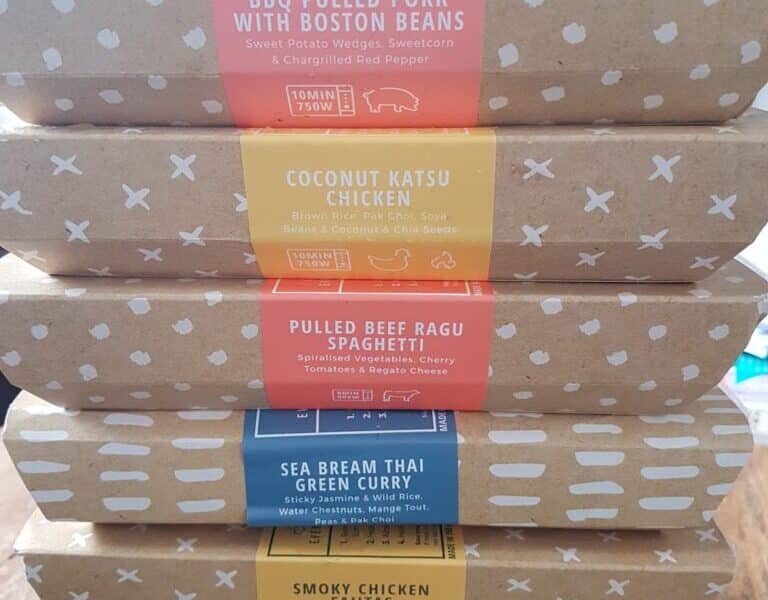 A stack of meal boxes with colourful labels, each describing a different dish, including Paneer & Sweet Potato Kofte Biryani, BBQ Pulled Pork with Boston Beans, Coconut Katsu Chicken, and more. The boxes are brown with a white pattern.