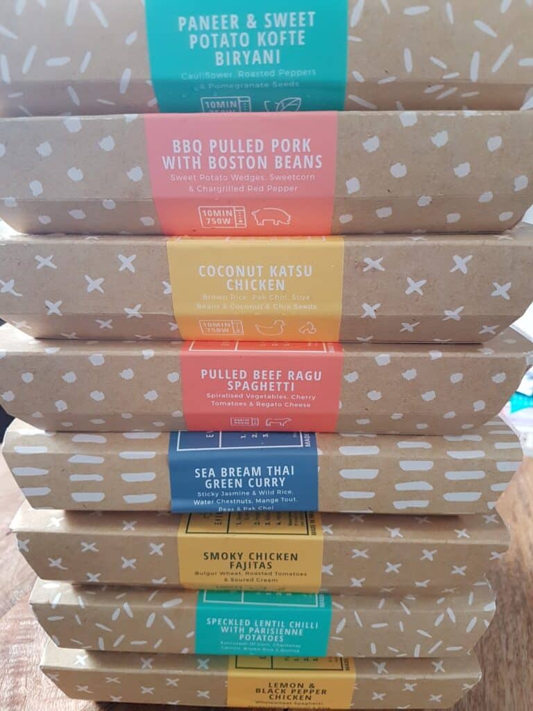 A stack of seven packaged ready meals, each labelled with a colourful strip describing the contents, including dishes such as Paneer & Sweet Potato Kofte Biryani, BBQ Pulled Pork, Coconut Katsu Chicken, and more, all in brown boxes with white patterns.