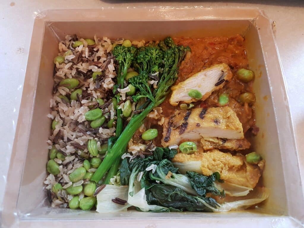 A meal in a plastic container with wild rice and edamame on the left, grilled chicken breast in an orange curry sauce with more edamame on the right, and steamed broccolini and pak choi arranged between the rice and chicken.
