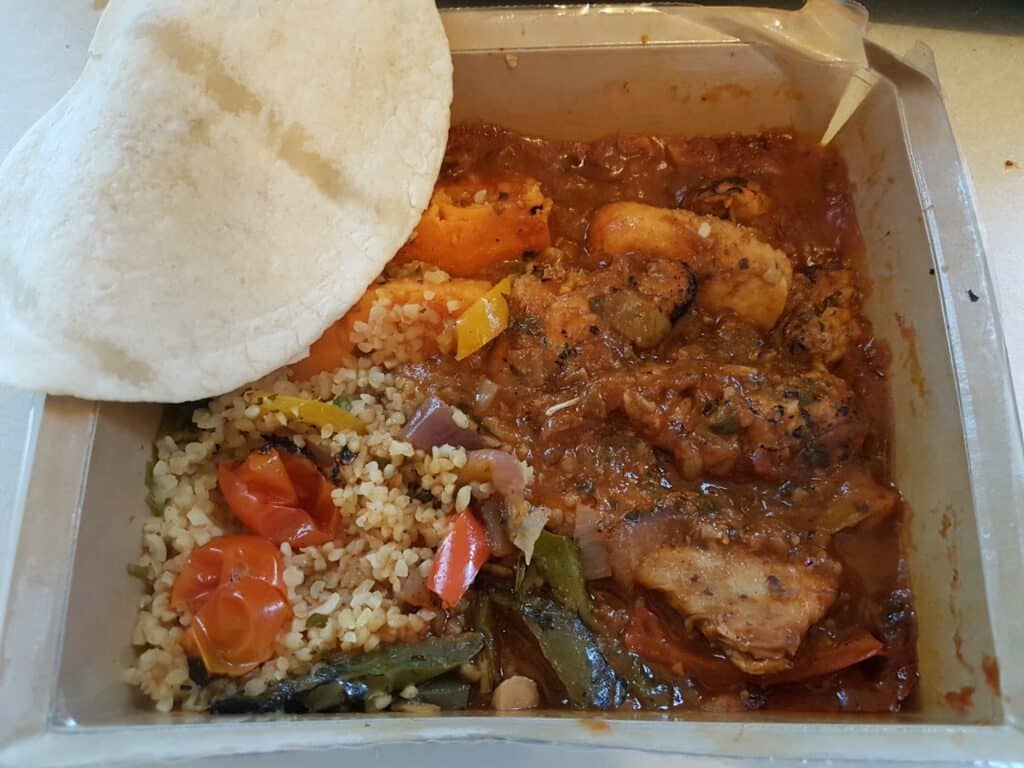 A meal in a plastic container features chicken in tomato-based sauce with herbs, bulgur mixed with grilled vegetables like cherry tomatoes, green beans, and onions, and is topped with a folded piece of flatbread.