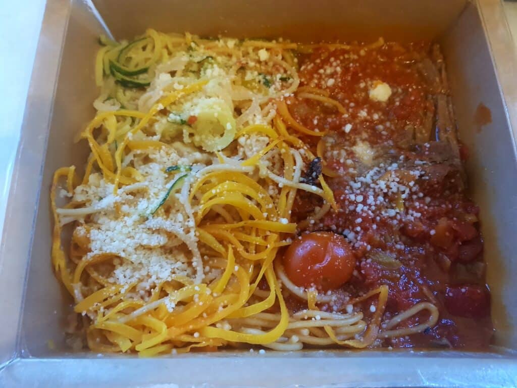 A divided takeaway container holds spiralised courgette, spiralised yellow squash, and spaghetti topped with tomato sauce, cherry tomatoes, grated parmesan, and herbs. The colourful vegetables and pasta are separated, sprinkled with cheese.