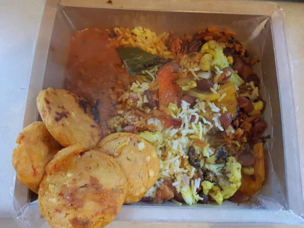 A plastic tray containing yellow and white rice mixed with vegetables like cauliflower and peas, a brown curry, and three round, golden-brown fried snacks placed on the side. The meal appears to be an Indian-style vegetarian dish.