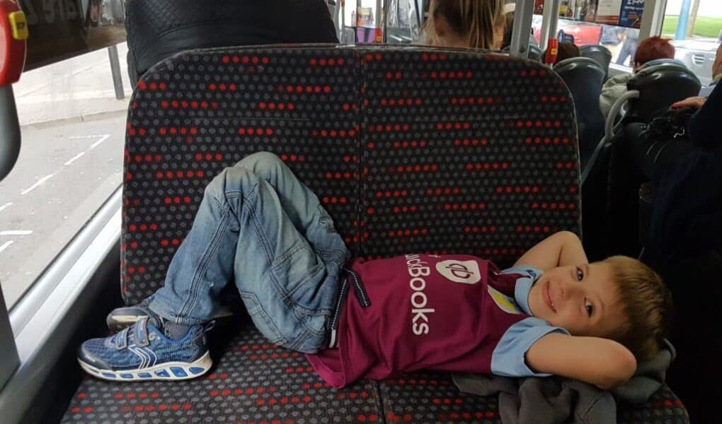 A young boy in jeans and a maroon shirt lies across a patterned bus seat with his hands behind his head, smiling at the camera. Other passengers sit nearby, and the view outside shows a coach and trees through the window.