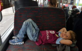 A young boy in jeans and a maroon shirt lies across a patterned bus seat with his hands behind his head, smiling at the camera. Other passengers sit nearby, and the view outside shows a coach and trees through the window.