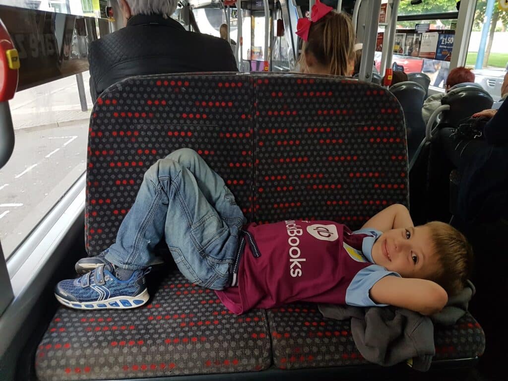 A young boy lies sideways on a patterned bus seat, legs stretched out and hands behind his head, smiling at the camera. He wears jeans, blue trainers, and a maroon sports jersey. Other passengers sit nearby, and a window shows a street and another bus outside.