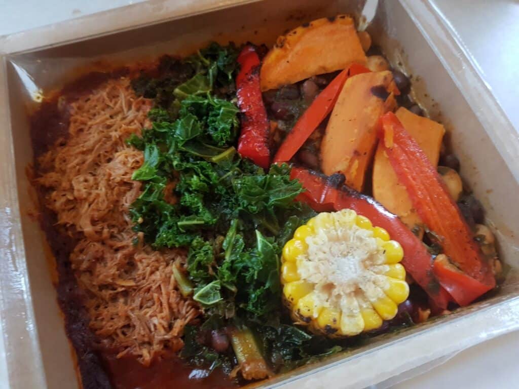A meal tray with shredded chicken in sauce on one side, and colourful vegetables on the other: sautéed kale, red pepper strips, sweet potato wedges, black beans, chickpeas, and a round slice of corn on the cob, all arranged neatly.