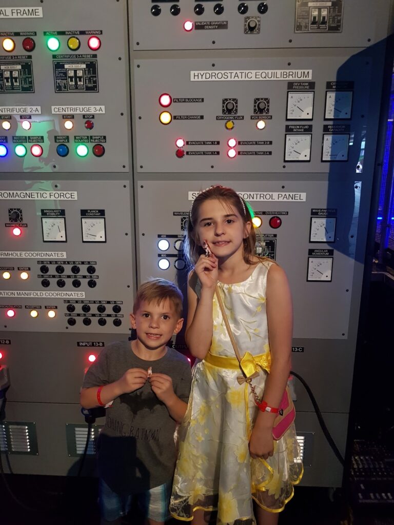 Two children stand in front of a large wall of scientific control panels with colourful buttons and switches. The girl wears a white dress with yellow flowers and holds a lollipop, whilst the boy wears a grey t-shirt and shorts, also holding a lollipop. Both are smiling.