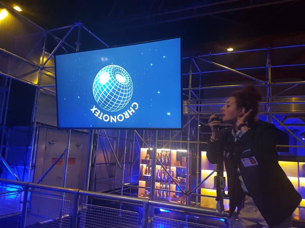 A woman with a microphone stands in front of a large screen displaying a blue globe and the white text "CHRONOTEX." The area is lit with blue light, featuring metal railings and scaffolding, and shelves with items are visible in the background.