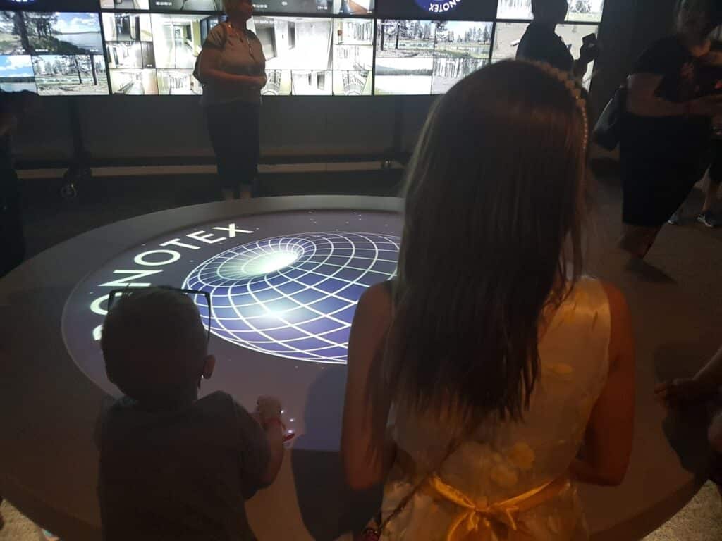 A girl in a white dress with a yellow sash and a young boy stand at a circular interactive table displaying a glowing globe and the word "CONOTEX." Several people are in the dimly lit background, with screens lining the back wall.