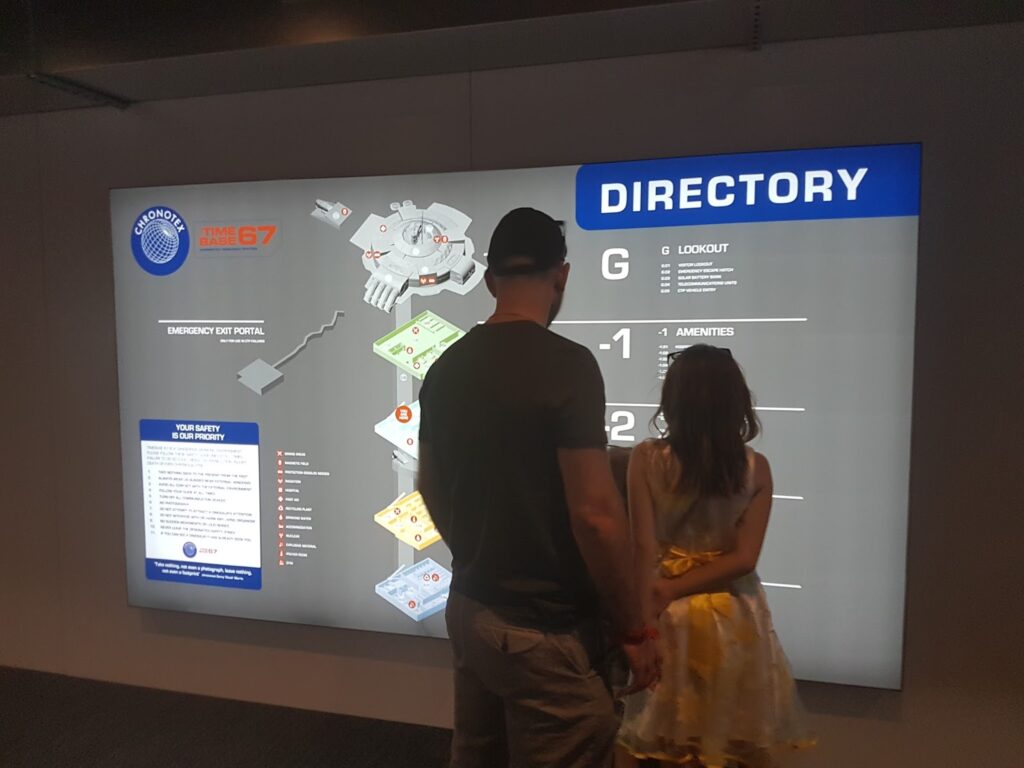A man in a black shirt and cap stands beside a young girl in a yellow dress, both facing and reading a large, illuminated building directory sign mounted on a wall indoors. The sign features maps, labels, and colour-coded sections.