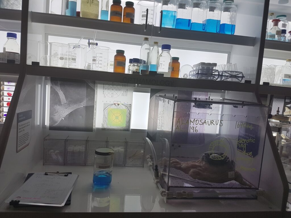 A laboratory scene with shelves of glass bottles, jars, and test tubes, labelled “OPHTHALMOLOGY.” A transparent enclosure holds a specimen labelled “MOSASAURUS.” A blue liquid is in a jar; a clipboard with papers lies on the counter. Scientific diagrams are on the wall.