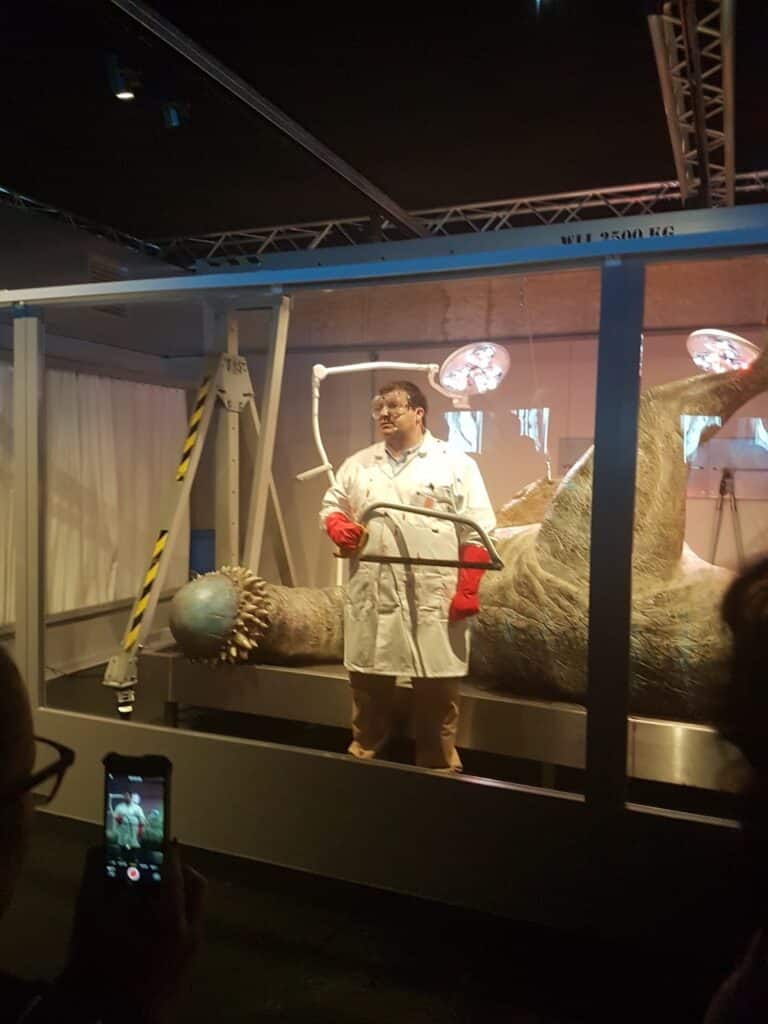 A person in a white lab coat and red gloves stands in front of a large dinosaur model on an examination table, holding a tool. Overhead lights shine down, and someone in the foreground is photographing the scene with a mobile phone.