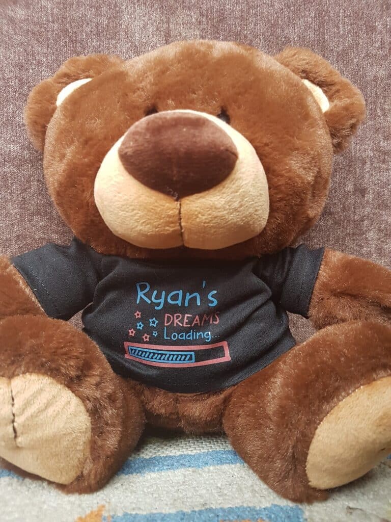 A brown teddy bear sits on a greyish-brown surface, wearing a black T-shirt that reads "Ryan's DREAMS Loading..." with blue and pink text and a loading bar graphic beneath the words. The bear’s face is centred, arms open, and feet visible.