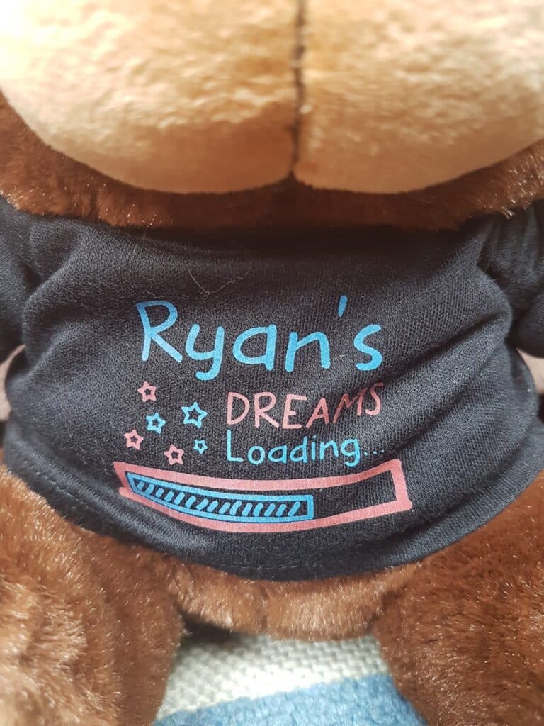 A brown stuffed toy wears a black shirt that reads “Ryan’s DREAMS Loading...” in blue and pink text, with stars and a colourful loading bar graphic below the words. The toy is shown from the neck down, focusing on the shirt's design.