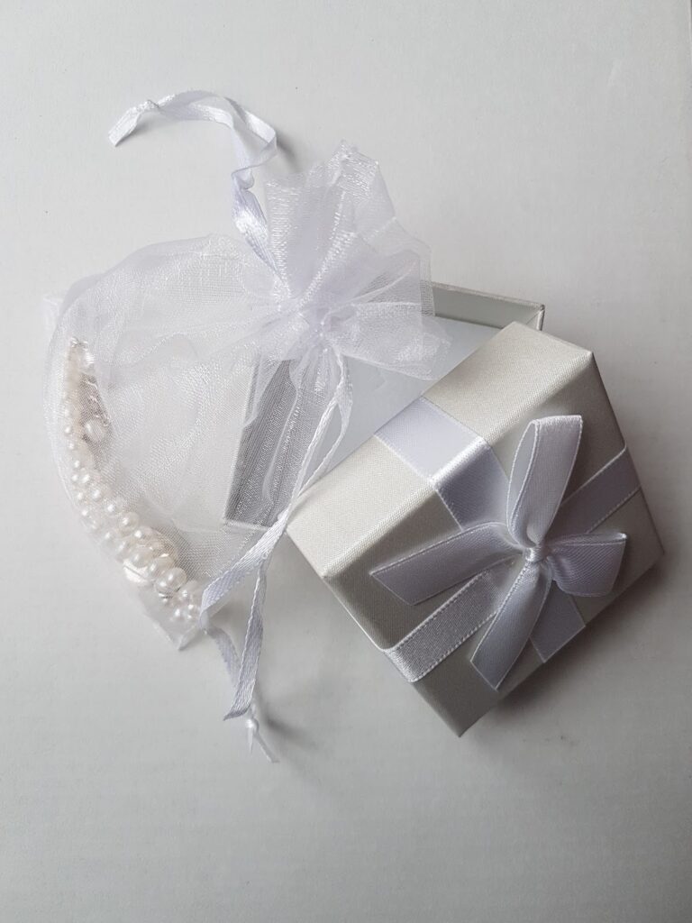 A white jewellery box with a white ribbon bow is open, revealing a sheer white drawstring pouch inside. The pouch contains a delicate pearl bracelet and a small rectangular item, all set on a plain white background.