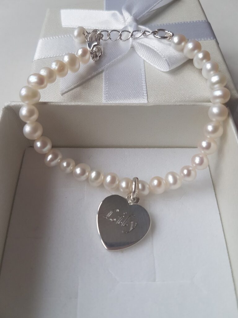 A pearl bracelet with a silver heart-shaped charm engraved with "Baby" is displayed in a white box with a white ribbon bow on the lid. The pearls are round and glossy, and the heart charm hangs from the centre of the bracelet.