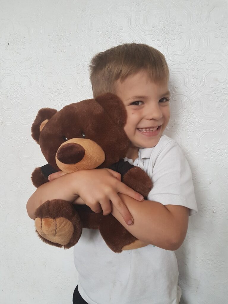A young child with short brown hair, wearing a white shirt, is smiling and hugging a large brown teddy bear. The background is a textured white wall with raised patterns. The child looks happy and content whilst holding the plush toy.