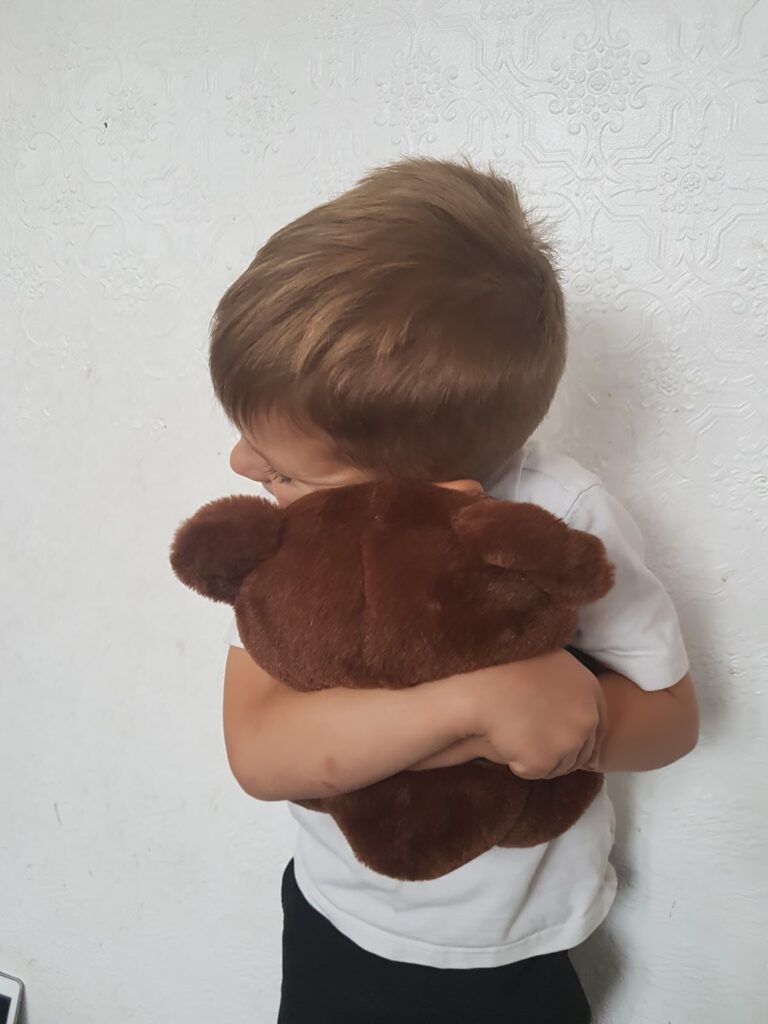 A young child with light brown hair, wearing a white shirt, hugs a brown teddy bear tightly whilst facing sideways against a textured white wall. The child’s face is partially hidden behind the teddy bear.