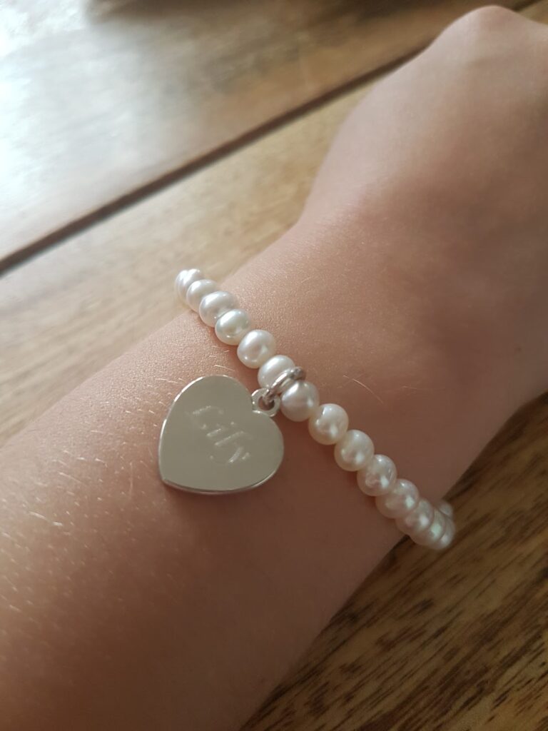 A close-up of a wrist wearing a bracelet made of white pearls with a silver heart-shaped charm. The charm has engraved text and is attached near the clasp. The background is a wooden surface.