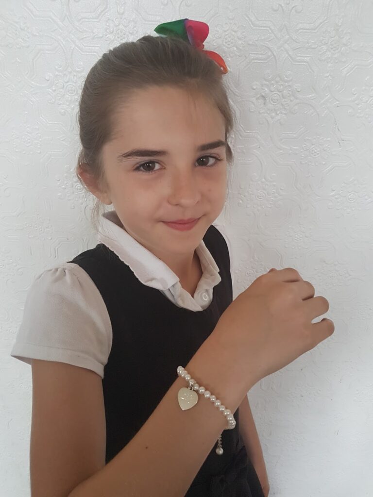A young girl with brown hair tied back and a multicoloured bow stands against a white textured wall. She wears a white collared shirt under a dark dress and smiles softly. On her wrist is a pearl bracelet with a gold heart charm.