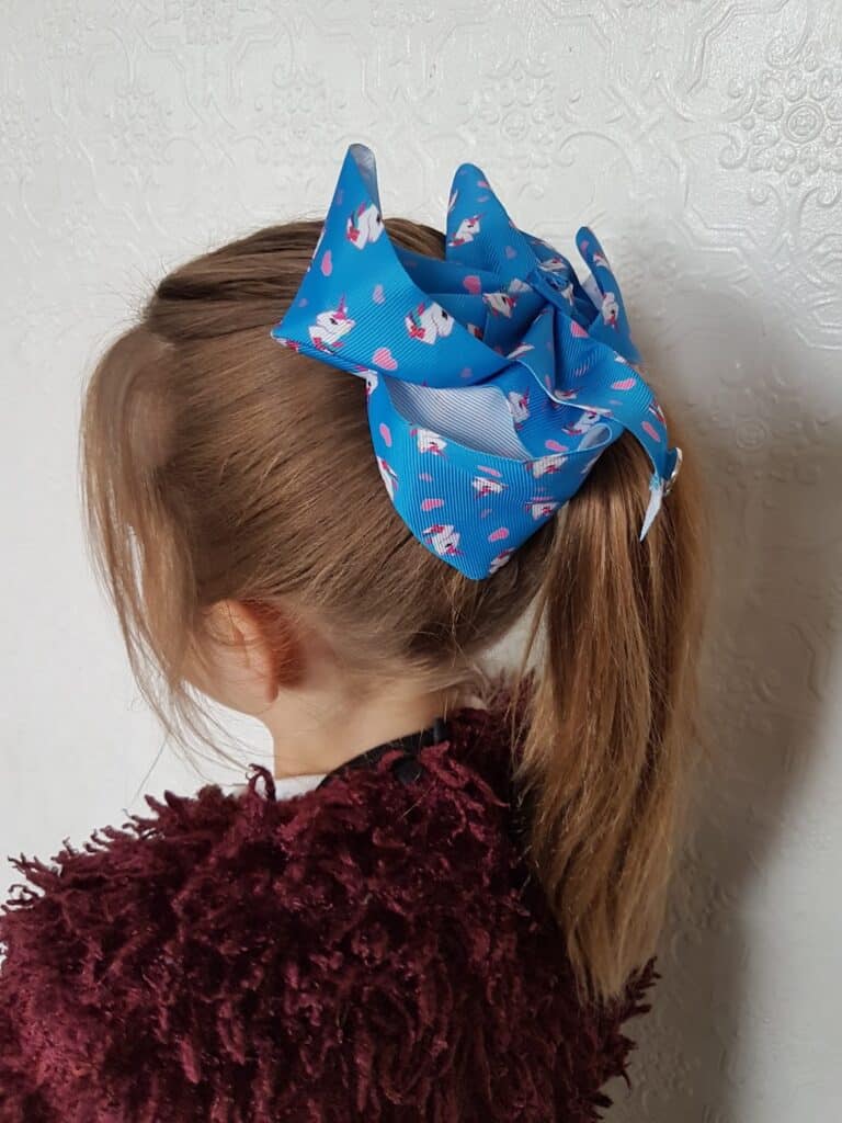 A young girl with light brown hair in a ponytail wears a large blue bow with pink and white unicorn designs. She’s dressed in a burgundy textured jumper, and the background is a white, patterned wall. The girl’s face is turned away from the camera.