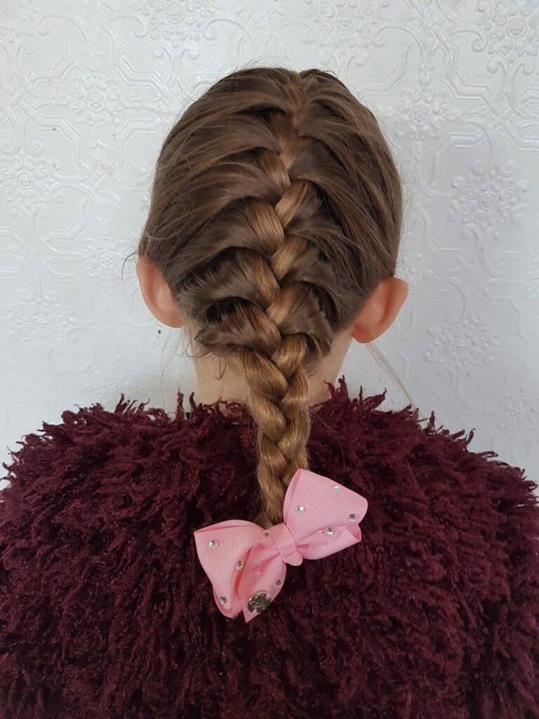 A girl with light brown hair styled in a neat French plait. She is wearing a fluffy maroon jumper and has a large pink bow with small white pearls at the end of her plait. The background is a white textured wall.