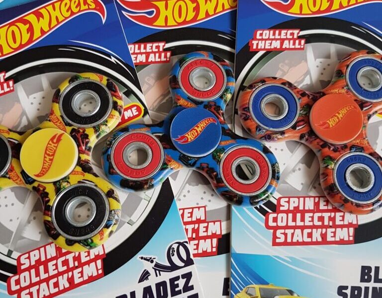 Three Hot Wheels-branded Bladez Spinnerz fidget spinners in packaging are shown. Each pack features a yellow sports car image, Hot Wheels logos, and colourful spinners with car-themed designs on a blue and white background.